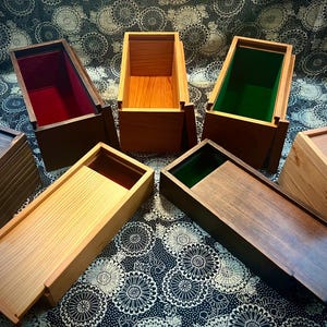 May include: A collection of rectangular wooden boxes with sliding lids, featuring various wood tones including light, medium, and dark finishes. Some boxes have interior linings in red or green, while others show natural wood interiors.