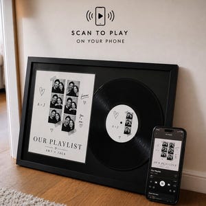 以下が含まれることがあります： 黒いフレームに入ったウォールアート。写真ストリップ、"OUR PLAYLIST"と"AMY + JACK"の文字、黒いレコードが白い台紙に配置されています。スマートフォンには同じプレイリストが表示され、上部には"SCAN TO PLAY ON YOUR PHONE"のアイコンがあります。