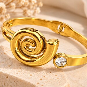 May include: A gold-toned hinged bangle bracelet featuring a prominent spiral design on the front and a single round clear gemstone set in a circular bezel. The bracelet has a polished metallic finish and a secure clasp mechanism on the side.