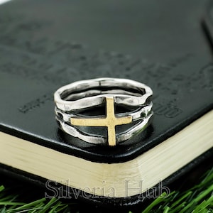 May include: A silver ring with a gold cross design. The ring has a layered band and a textured finish. The gold cross is centered on the ring. The ring is displayed on a black surface with the text "Silveria Hub".