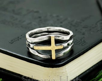 May include: A silver ring featuring a gold cross design. The ring has a layered band and a textured finish. The gold cross is centrally placed. The ring is shown on a black surface with the text "Silveria Hub".