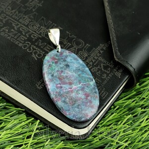 May include: Oval pendant with a silver bail. The pendant displays a mottled pattern of blue, green, and maroon colours. The gemstone is set against a black background, with a hint of green grass visible.