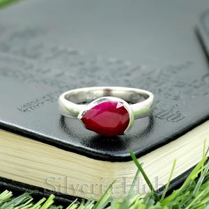 Puede incluir: Un anillo de plata con una piedra preciosa roja oscura en forma de lágrima. El anillo tiene una banda pulida y la piedra preciosa está engastada en un bisel. El anillo se muestra en un cuaderno negro.