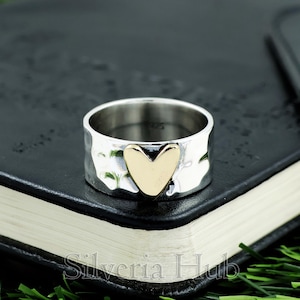 Op de afbeelding: Een zilveren ring met een gehamerde textuur en een gouden hartvormig accent. De ring is breed en ligt op een donkere ondergrond. De ring is een sieraad.