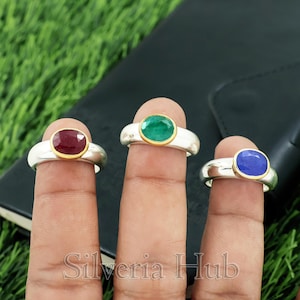 Può includere: Tre anelli con pietre preziose ovali in rosso rubino, verde smeraldo e blu zaffiro. Ogni anello ha una fascia argentata con una lunetta dorata attorno alla gemma. Gli anelli sono esposti su dita.