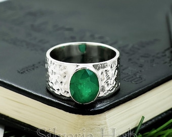 Puede incluir: Un anillo de plata con una banda texturizada y una piedra preciosa de esmeralda ovalada y facetada. La banda del anillo tiene un diseño martillado. La esmeralda es de color verde vibrante. El anillo se muestra sobre una superficie oscura.