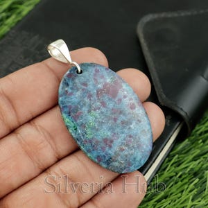 Puede incluir: Colgante ovalado con una fianza plateada. El colgante presenta una piedra azul y granate con detalles verdes. El texto "Silveria Hub" es visible. Probablemente sea una pieza de joyería.