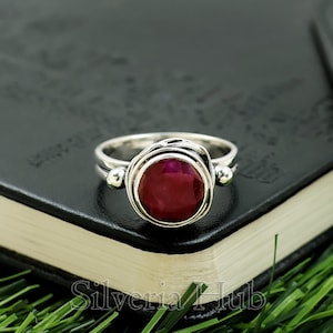 May include: A silver ring featuring a large, round, deep red gemstone. The ring has a distinctive design with a double band and decorative details on the sides. The ring is set against a dark background.