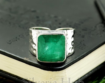 Puede incluir: Un anillo de plata con una gran piedra preciosa verde esmeralda cuadrada. El anillo tiene una banda texturizada y un diseño abierto. La piedra preciosa es el punto focal, mostrando su color y corte vibrantes.