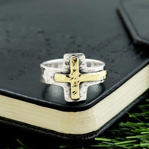 May include: A silver ring with a gold cross design. The ring has a textured band and a hammered finish on the cross. The gold cross is centered on the ring, creating a bold and religious statement. The ring is set against a dark background.