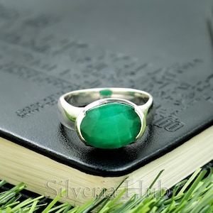 Puede incluir: Un anillo de plata con una gran esmeralda de talla ovalada. La piedra verde brillante está engastada en una banda de plata pulida. El anillo se muestra sobre una superficie negra con el texto "Silveria Hub" visible.