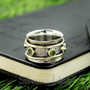 May include: A silver and gold-tone spinner ring with three green gemstones. The ring has a textured band and a rotating top. The ring is displayed on a dark surface with the text "Silveria Hub".