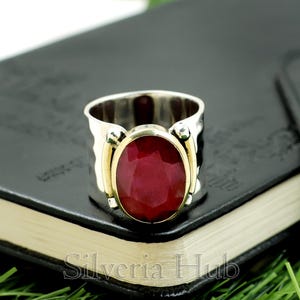 Può includere: Un anello d'argento con una grande gemma ovale di colore rosso intenso. La gemma è incastonata in una lunetta color oro e l'anello ha una fascia larga. L'anello è esposto su una superficie nera.