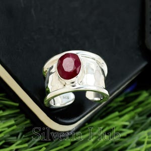 Puede incluir: Un anillo ancho de plata con una textura martillada y una gran gema ovalada de rubí. El anillo tiene un diseño abierto y está colocado sobre un fondo oscuro. El texto "Silveria Hub" es visible en la parte inferior.