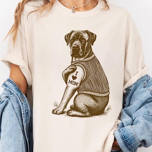 May include: A cream-coloured t-shirt with a brown illustration of a dog. The dog has a tattoo that reads "I ❤️ MOM" and wears a striped sweater. The shirt has short sleeves and a relaxed fit.