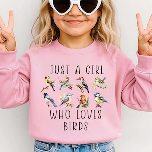 May include: Pink sweatshirt with the text "JUST A GIRL WHO LOVES BIRDS" and a colorful bird graphic. The sweatshirt is worn by a person wearing heart-shaped sunglasses and making a peace sign.