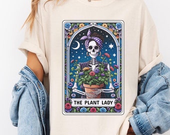 Puede incluir: Una camiseta blanca con un gráfico de un esqueleto sosteniendo una planta en maceta. El gráfico está en un estilo de tarjeta de tarot vintage con el texto "THE PLANT LADY".