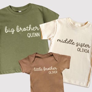 May include: Three children's shirts and onesie. The olive green t-shirt says "big brother QUINN". The cream t-shirt says "middle sister OLIVIA". The brown onesie says "little brother OLIVER".