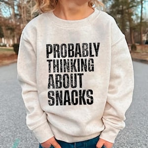 Pode incluir: Camisola bege com o texto "PROBABLY THINKING ABOUT SNACKS" em preto. A parte superior de manga comprida e gola redonda é usada por uma criança. Calças de ganga azuis são visíveis na parte inferior.