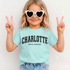 May include: A light blue t-shirt with the word "CHARLOTTE" in black block letters and "NORTH CAROLINA" underneath. The person is wearing white heart-shaped sunglasses and making a peace sign with both hands.