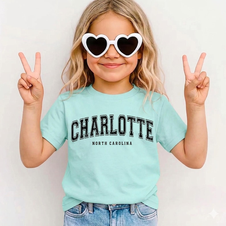 May include: A light blue t-shirt with the word "CHARLOTTE" in black block letters and "NORTH CAROLINA" underneath. The person is wearing white heart-shaped sunglasses and making a peace sign with both hands.