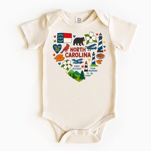 May include: Cream-colored baby bodysuit with a heart-shaped graphic featuring North Carolina state symbols. The design includes a flag, bear, flowers, lighthouse, and text that reads "North Carolina" and "First in Flight."