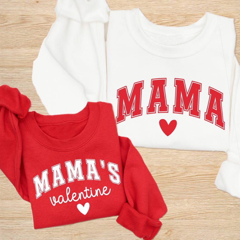 May include: Two sweatshirts, one red and one white. The red sweatshirt has the text "MAMA'S valentine" in white, with a heart. The white sweatshirt has the text "MAMA" in red, with a heart. Both are crew neck style.