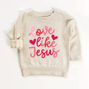 Peut inclure: Un sweat-shirt beige avec le texte "love like Jesus" en lettres roses et rouges avec des c&oelig;urs.