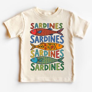 May include: Cream-colored t-shirt featuring the word "SARDINES" above three colorful fish illustrations. The fish are red, orange, and blue, each with unique patterns and details. The text and fish are outlined in black.