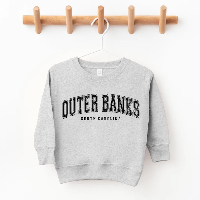 May include: A light gray sweatshirt with the text "OUTER BANKS NORTH CAROLINA" in black, hanging on a wooden rack. The sweatshirt has a crew neck and long sleeves, suitable for children.