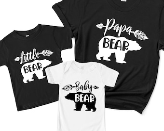 May include: Three black t-shirts with white bear silhouettes and text. The largest shirt says "Papa Bear" with an arrow and feather design. The medium shirt says "Little Bear" with an arrow and feather design. The smallest shirt says "Baby Bear" with an arrow and feather design.