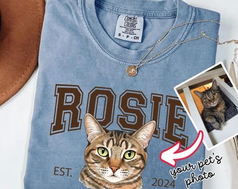 May include: A blue Comfort Colors t-shirt featuring a custom cat portrait and the name "ROSIE" in brown collegiate lettering. The shirt includes the text "EST. 2024" and a small photo of a cat. A gold necklace is draped across the shirt.