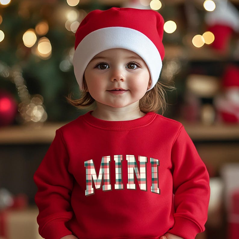 May include: A child wearing a red Santa hat with a white brim and a red sweatshirt. The sweatshirt has the word "MINI" in a plaid design across the front. The background is blurred with Christmas lights.