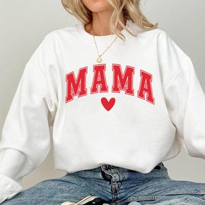 May include: White sweatshirt with the red word "MAMA" in a collegiate font and a red heart below. The sweatshirt is a crew neck style.
