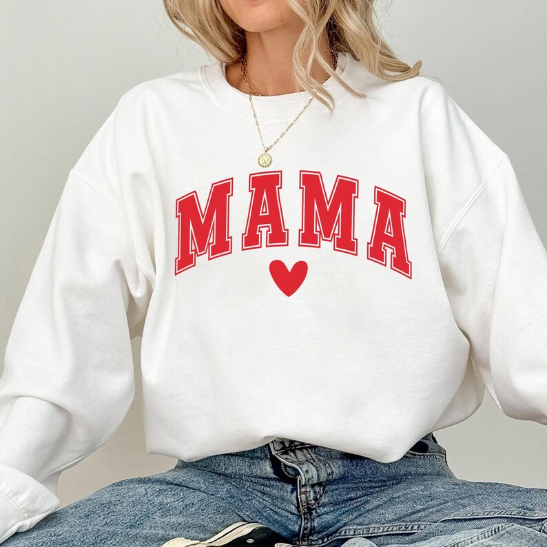 May include: White sweatshirt with the red word "MAMA" in a collegiate font and a red heart below. The sweatshirt is a crew neck style.