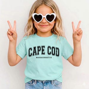 May include: A young person wearing heart-shaped sunglasses and a light blue t-shirt with the words "CAPE COD MASSACHUSETTS" printed in black. The person is giving a peace sign with both hands.