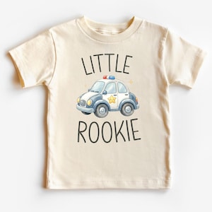 May include: Cream-colored toddler t-shirt with the words "LITTLE ROOKIE" in black. A cartoon police car with a star on the side is in the center. The car is white, blue, and gray with red and blue lights on top.