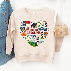 May include: A beige sweatshirt featuring a heart-shaped design with North Carolina state symbols. The design includes a bear, lighthouse, airplane, and the state flag. Text includes "North Carolina", "First in Flight", and "From Mountains to Coast".