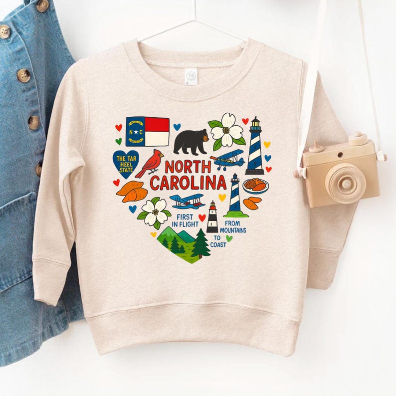 May include: A beige sweatshirt featuring a heart-shaped design with North Carolina state symbols. The design includes a bear, lighthouse, airplane, and the state flag. Text includes "North Carolina", "First in Flight", and "From Mountains to Coast".