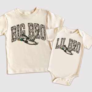 May include: Two matching cream-colored shirts. One is a t-shirt with "BIG BRO" in camouflage lettering and a flying duck graphic. The other is a baby onesie with "LIL BRO" in similar lettering and a duck graphic.