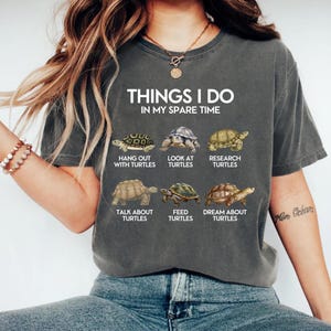 Pode incluir: Uma camiseta cinza escuro com o texto "THINGS I DO IN MY SPARE TIME" e ilustrações de tartarugas. A camiseta lista atividades como "Hang out with turtles" e "Dream about turtles".