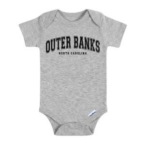 May include: A gray baby bodysuit with short sleeves. The text "OUTER BANKS" is arched across the top in black letters with a white outline, and "NORTH CAROLINA" is below. The bodysuit has a snap closure.