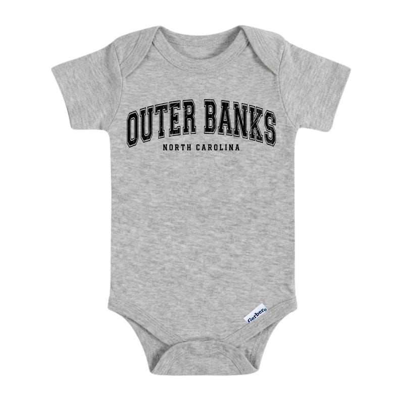 May include: A gray baby bodysuit with short sleeves. The text "OUTER BANKS" is arched across the top in black letters with a white outline, and "NORTH CAROLINA" is below. The bodysuit has a snap closure.
