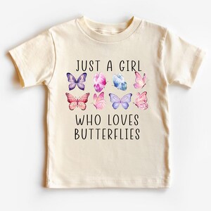 May include: Cream-colored t-shirt featuring the text "JUST A GIRL WHO LOVES BUTTERFLIES" in black. The shirt has watercolor illustrations of colorful butterflies in shades of purple, pink, and blue.