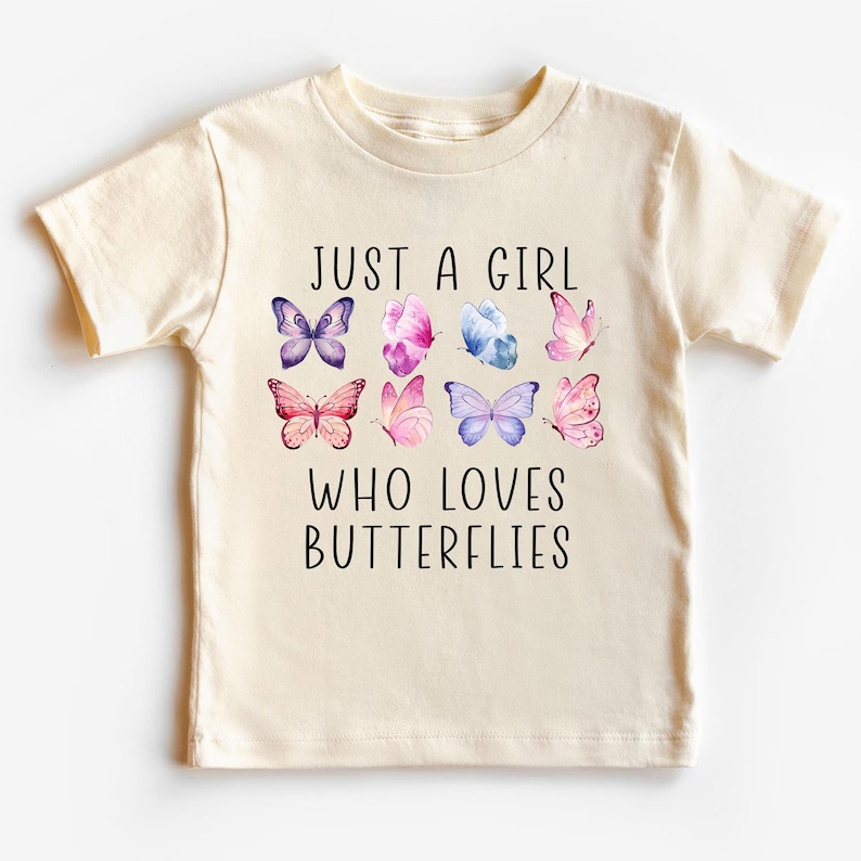 May include: Cream-colored t-shirt featuring the text "JUST A GIRL WHO LOVES BUTTERFLIES" in black. The shirt has watercolor illustrations of colorful butterflies in shades of purple, pink, and blue.