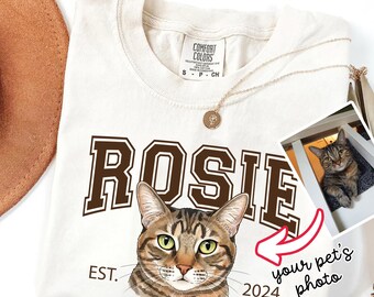 May include: Cream-colored t-shirt with the name "Rosie" in brown block letters and a detailed cat illustration. The shirt includes the text "EST. 2024" and a small photo of a cat. A gold necklace and a brown hat are also visible.