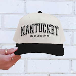 Può includere: Un cappellino da baseball color crema con visiera nera e bottone nero sulla parte superiore. Il cappellino presenta la scritta "NANTUCKET" in grassetto nero sopra "MASSACHUSETTS".