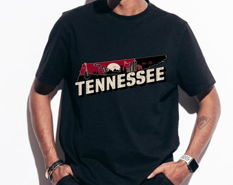 Puede incluir: Camiseta negra con un diseño de Tennessee. El diseño incluye la silueta del estado con un paisaje urbano, una guitarra y la palabra "Tennessee" en una fuente vintage. La camiseta es de manga corta.