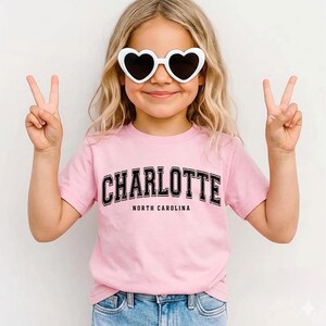 May include: A young person wearing a pink t-shirt with the word "CHARLOTTE" in black block letters, with "NORTH CAROLINA" below. They are also wearing white heart-shaped sunglasses and making a peace sign with both hands.