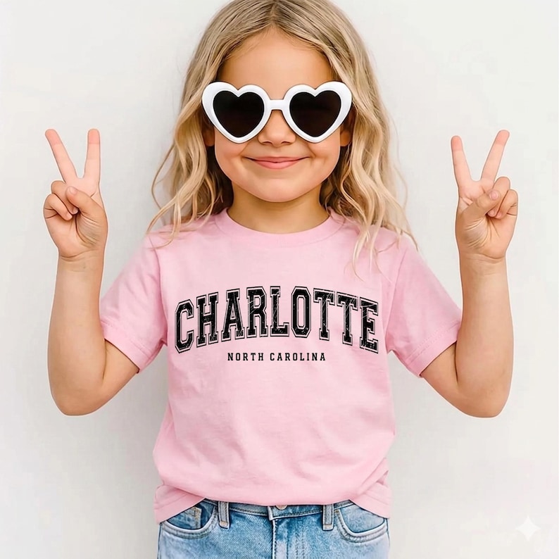 May include: A young person wearing a pink t-shirt with the word "CHARLOTTE" in black block letters, with "NORTH CAROLINA" below. They are also wearing white heart-shaped sunglasses and making a peace sign with both hands.